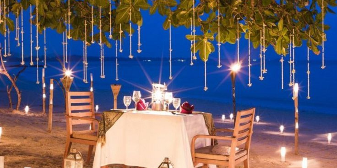 A romantic, intimate dinner setting for two on the sand beneath a canopy of illuminated foliage against a deep twilight sky.