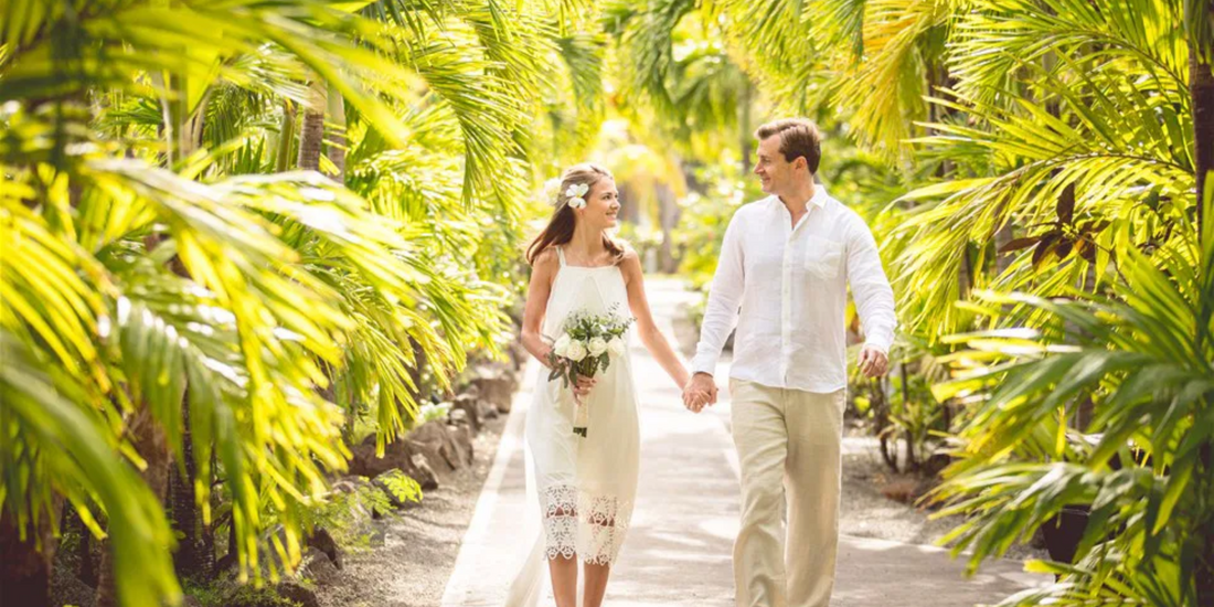 A couple walks hand-in-hand down a paved path, surrounded by lush tropical foliage.