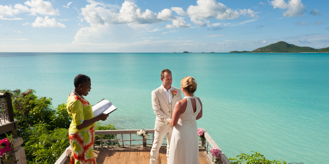 A couple exchanges vows on a wooden platform overlooking a turquoise ocean, bathed in sunlight.