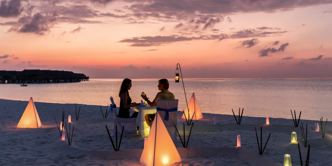 A couple enjoys a private, candlelit dinner on a white sand beach at sunset, with overwater bungalows visible in the distance.