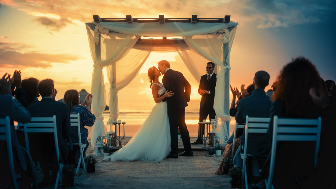 Guests cheer as the bride and groom exchange their first kiss at their beach wedding ceremony, bathed in the golden light of sunset.