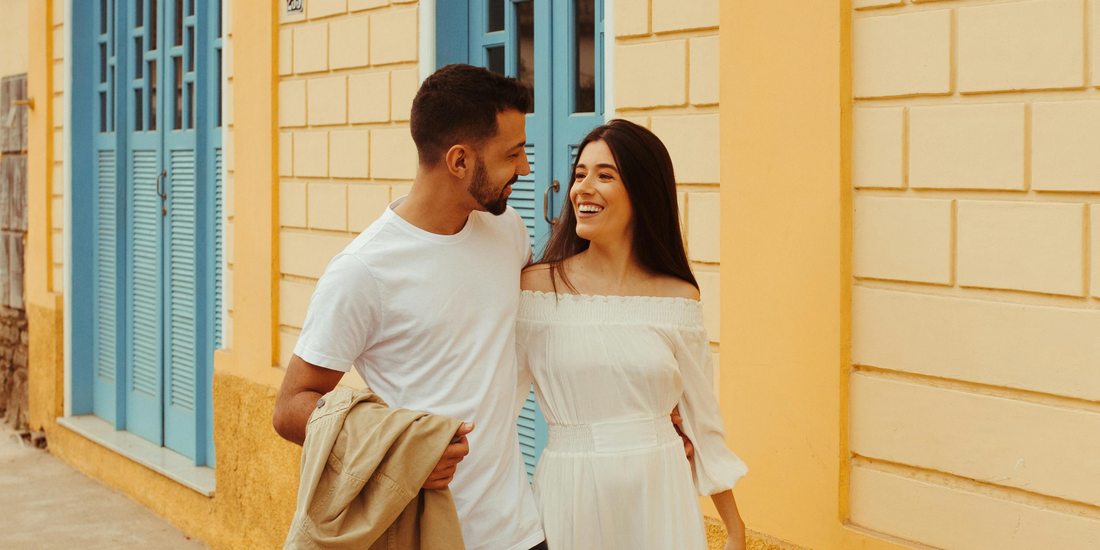 A couple walking outdoors against a bright yellow, textured wall with light blue shutters.