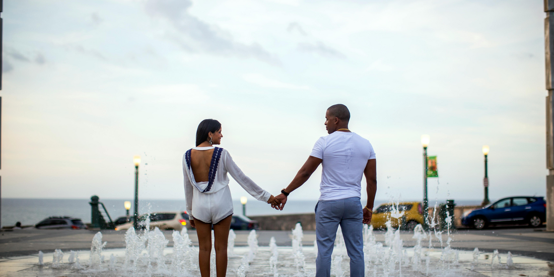 Couple holding hands while standing near a public fountain with the ocean visible in the background during overcast daylight.