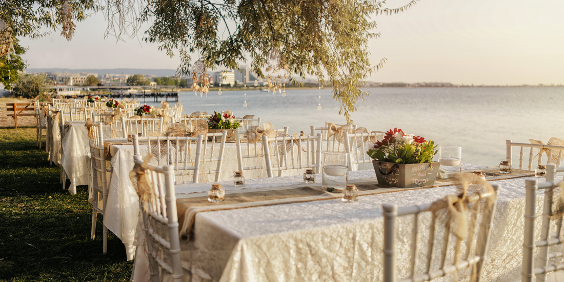 Tables set for an event are elegantly arranged outdoors by the water, with white chairs and floral centerpieces creating a charming atmosphere.