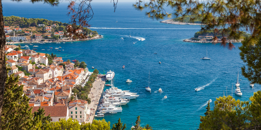 Aerial view capturing the terracotta rooftops of a coastal town bordering vibrant, deep blue Adriatic waters filled with docked and moving vessels.