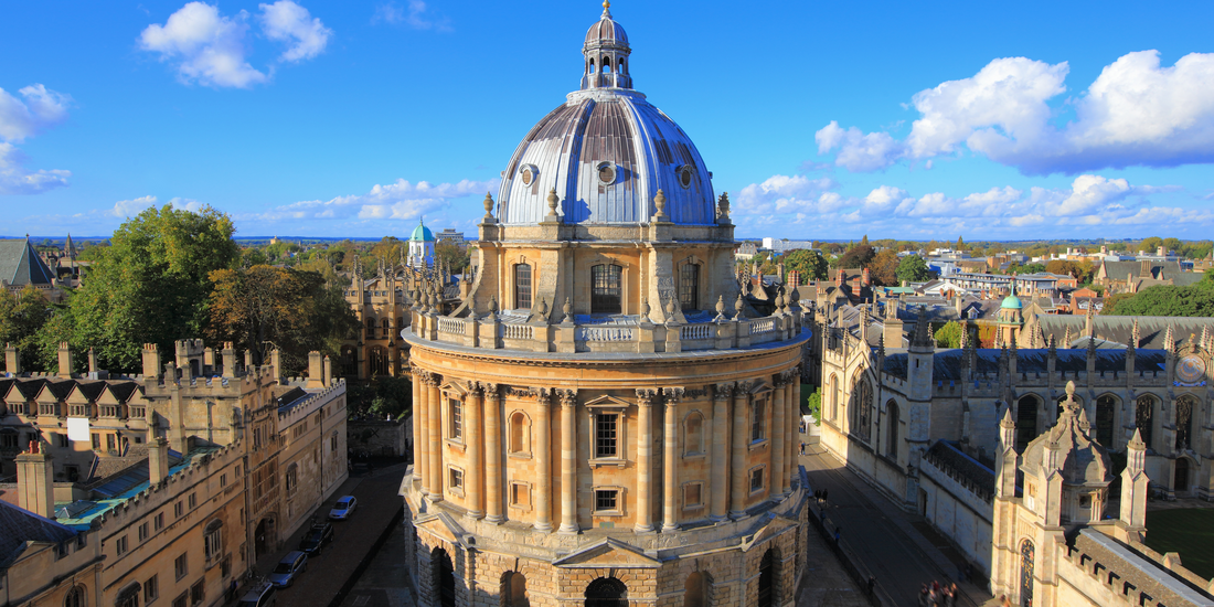 Aerial view capturing the Radcliffe Camera, an iconic circular library building at the University of Oxford, set against a bright blue sky with scattered white clouds.