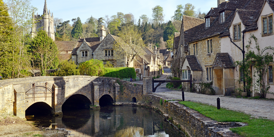 Stone bridge with multiple arches spanning a narrow waterway alongside traditional Cotswold stone cottages in an English village setting.