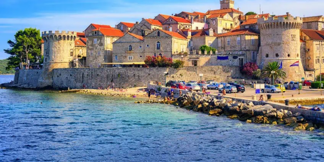 Coastal town featuring historic stone fortifications, terracotta rooftops, and a small beach area adjacent to the turquoise sea.