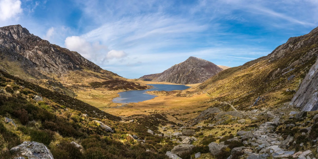Panoramic view of a rugged mountain valley showcasing a dark blue lake nestled between rocky slopes under a bright, partly cloudy sky.