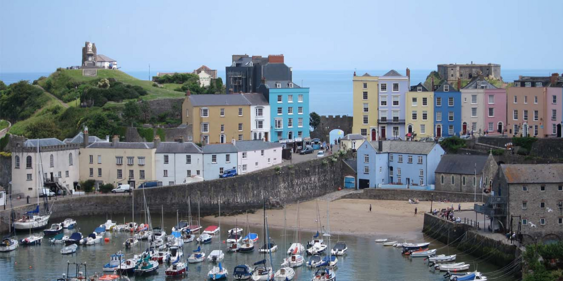 Sailboats moored in the foreground of a coastal settlement, with a sandy beach visible behind the quay wall.