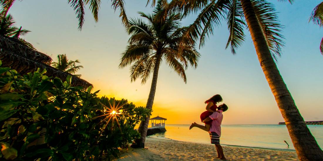 A couple embraces on a sandy tropical beach during a vibrant sunset, framed by tall palm trees and lush foliage.