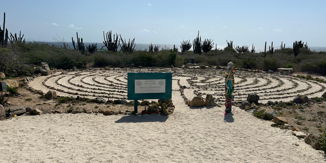 Outdoor labyrinth in Aruba, constructed from stones on a sandy path, set against a backdrop of arid coastal vegetation and the ocean.
