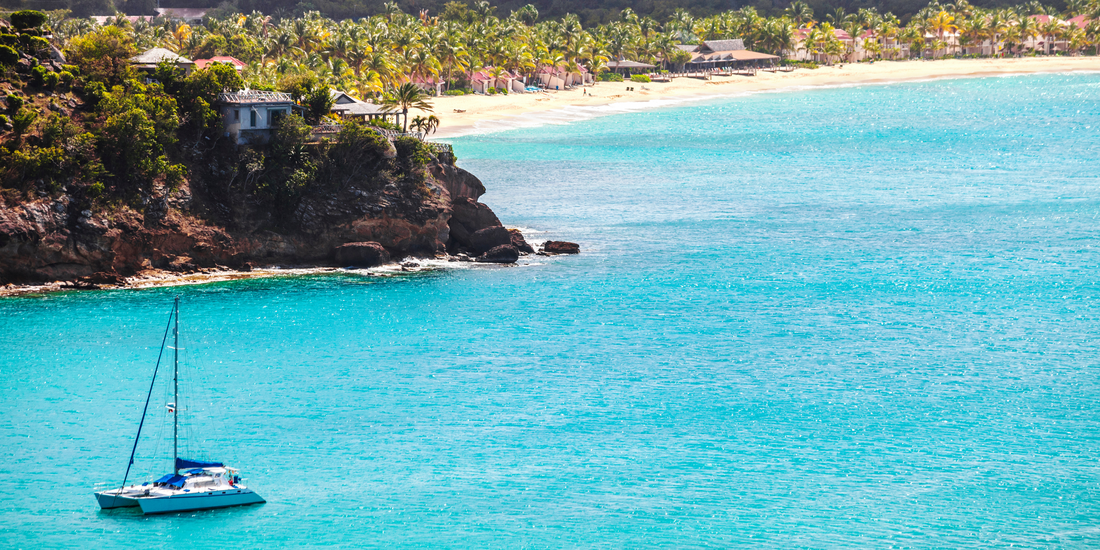 A catamaran rests on vibrant turquoise water near a rugged, vegetation-covered cliff face overlooking a distant sandy beach lined with palm trees.