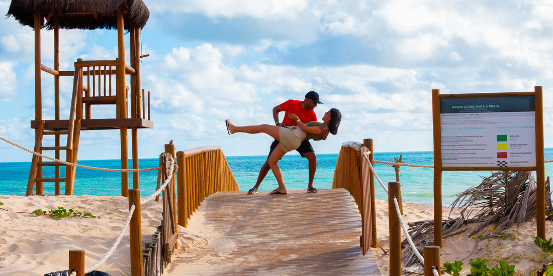 A couple performs a dance dip on a wooden boardwalk leading to a bright blue ocean, with a thatched lifeguard stand visible on the left.