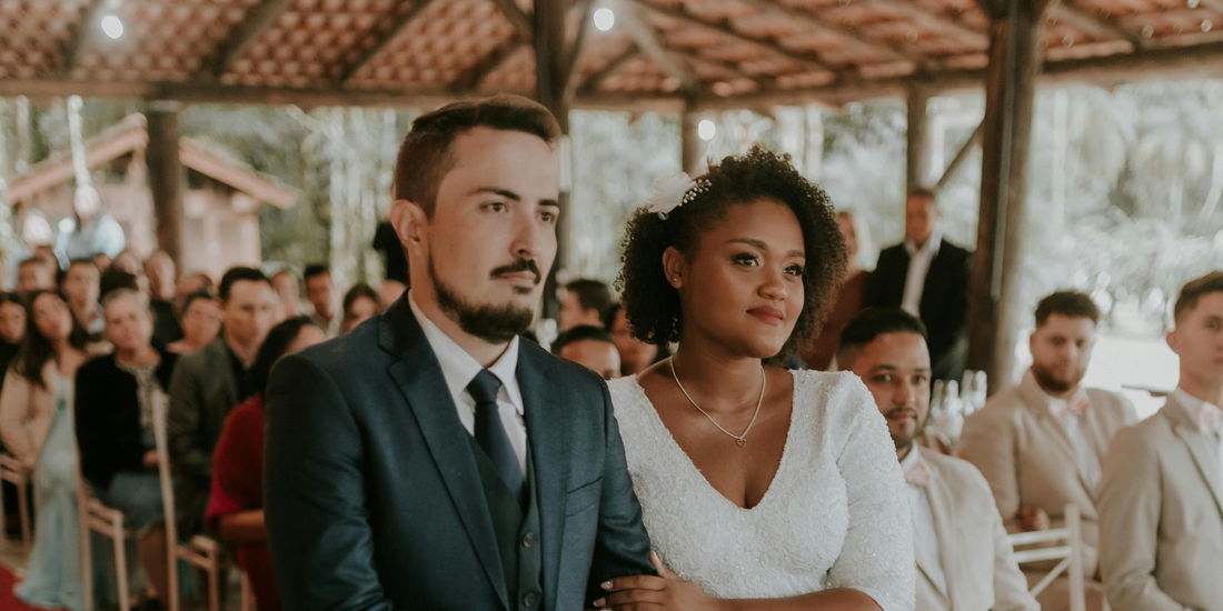 The bride and groom stand together during their wedding ceremony, surrounded by seated guests under a covered, open-air venue.