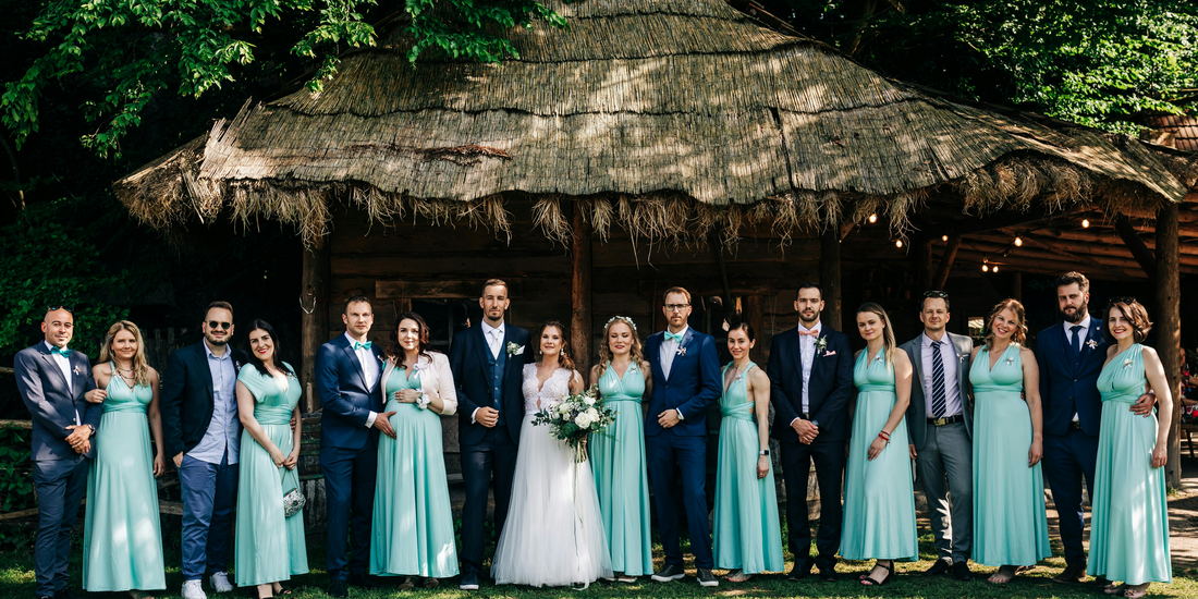 Full wedding party posing outdoors in front of a rustic, thatched-roof wooden structure.