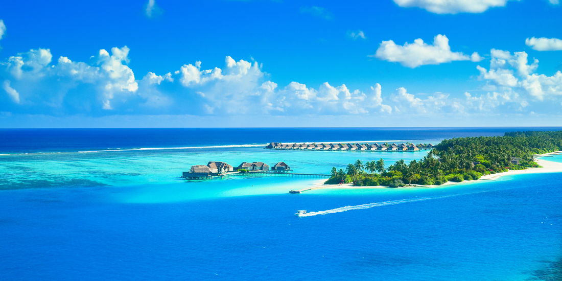 Overwater cottages and a tropical island shoreline in the Cayman Islands surrounded by vivid blue water and lush palm covered land.