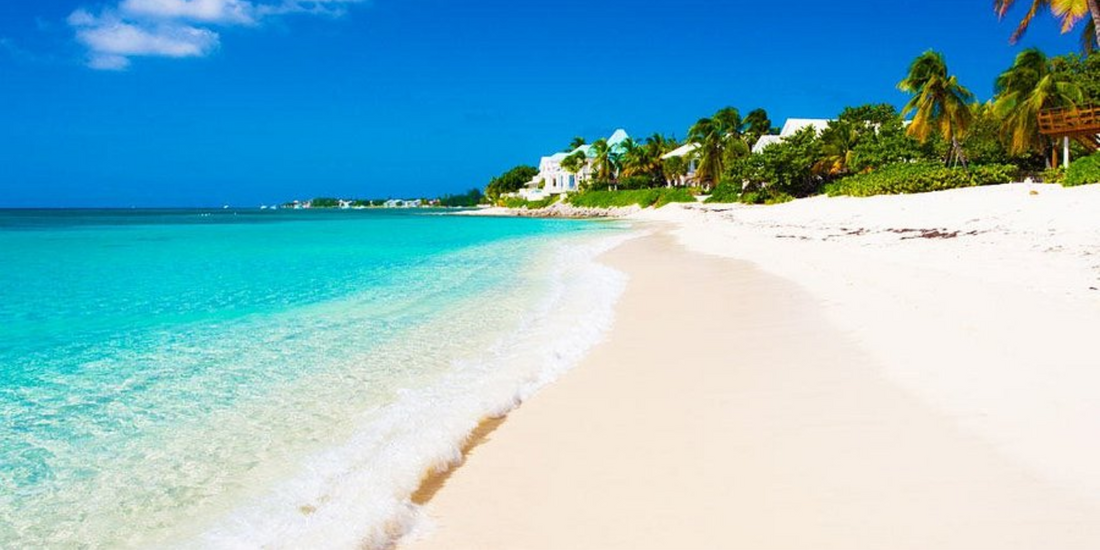 Sunny day along Seven Mile Beach in the Cayman Islands with soft white sand, turquoise water, palm trees, and coastal homes in the distance.