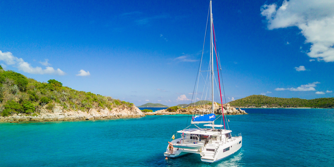 White sailing yacht anchored in calm turquoise waters of the British Virgin Islands, framed by rocky coastline, green hills, and clear blue skies.
